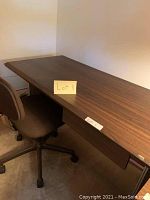 Woodgrain desk with brown fabric swivel chair against wall, photo showing lot sign with number 1 on desk
