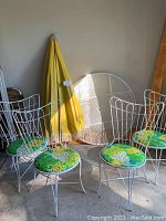 Four white metal outdoor chairs with curved wire backs and floral green-yellow cushions, yellow umbrella folded on side, round white metal mesh table top leaning against wall.