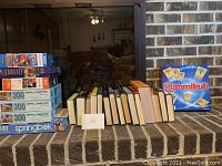 Arrangement showing seven puzzles with counts from 300 to 1000 pieces, fifteen assorted books lined up, and Rummikub game box on the side.
