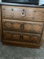 Front view of a wooden cabinet with three drawers featuring metal ring pulls and decorative metal studs.