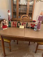 Overview of wooden Christmas decorations displayed on a wooden table, includes two wooden angels, two Santas, a snowman, and 'NOEL' letters.