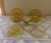 Five pieces of amber depression glassware on lace cloth, showing various patterns and shapes including round and square plates and a footed bowl.