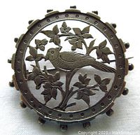 Front view of round antique sterling silver brooch with open work engraved bird and leaves design and cannonball indented border.