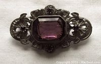 Front view of vintage white metal brooch with large faceted purple glass stone and ornate floral design