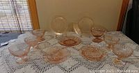 Overview of pink depression glass bowls, plates, and compote dishes arranged on a lace tablecloth, showing different patterns and sizes.