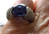 Close-up photo of the ring showing a faceted blue sapphire stone and pavé set clear stones on each shoulder.
