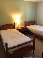 Two twin wooden beds with headboards and footboards side by side in a room, each with a mattress and a nightstand lamp between them.