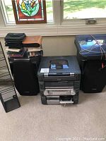 Photo showing Aiwa stereo system with speakers on either side, stacked CDs and cases on one speaker, and black metal CD stand in front.
