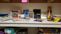 Wide photo showing a variety of craft-related items arranged on a shelf including books, paints, an embroidery thread pack, and paper plane instructions.