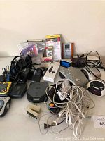 Lot of mixed used electronics including digital clocks, cassette players, portable players, earphones, extension cords, and plugs all arranged on table surface.