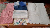 Overview of linens arranged together showing folded blue cotton tablecloth and napkins, embroidered white tablecloth and napkins, red and white apron, and various placemats.