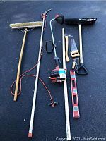Photo showing full view of various garden tools including push broom, weed wacker, tree looper, shovel, hand saw, axe, and bubble level laid out on a black surface.