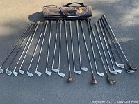 Photo shows a collection of golf clubs laid out on pavement with shadows, including multiple irons and wooden drivers, alongside a Proline golf bag.