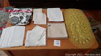 Various linens including folded tablecloths, napkins, boxed napkins, woven placemats, and apron arranged on wooden coffee table
