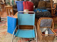 Full lot overview with blue lawn chair, wicker picnic basket with contents, red Coleman sleeping bag in compression sack, blue bucket/ice tub, black fishing rod, and black and blue sleeping bag