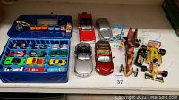 Overall display showing blue plastic storage case with miniature cars inside and additional larger vehicles and toys placed outside the case on a shelf.