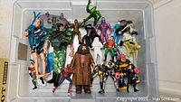 Top view of plastic bin filled with a mix of action figures including identifiable characters such as Spider-Man and a figure resembling Hagrid, as well as others in camouflage and various costumes.