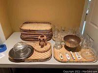 Multiple wicker trays stacked, various glass and plastic cups, metal bowl, glass dome cover, and bottle openers arranged on a white table.
