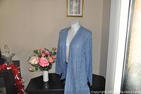 Light blue plus size open-front cardigan sweater displayed on mannequin near pink flowers and window blinds.