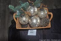 Basket holding 5 gold marbled glass ornaments with pine cones and greenery on a dark surface