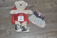 Build-A-Bear seated in red pet carrier backpack with adjustable straps and wearing black sneakers, next to the summer dress on the floor