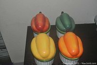 Top-down view of four bell pepper shaped ceramic serving dishes with lids in red, yellow, orange, and green colors on a wooden surface.
