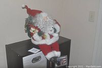 Photo showing plush Santa figurine sitting on a black cabinet shelf, dressed in red and white, holding gifts and a white bear, illustrating the sitting posture and festive details.