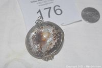 Close-up of the abalone shell pendant showing the shell pattern and metallic frame with clasp, placed next to a quarter for size reference.