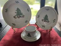 Display of a dinner plate, salad/dessert plate, cup, and saucer from the set showing Christmas tree and holly motifs with gold trim on a red cloth background.