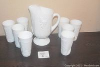 Set of one milk glass pitcher and eight tumblers with grape cluster embossed design, arranged on a table