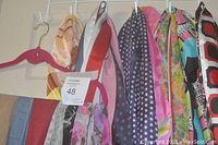 Twelve ladies scarves hanging on hangers on a wall rack and door. Various colors and patterns visible including solids and prints.