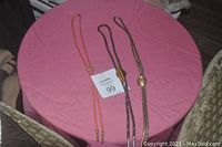 Lot of three men's western style bolo ties on a pink round tablecloth, showing orange, brown, and black braided strands with distinct fasteners.