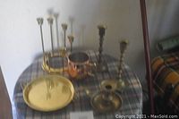 Photo showing a 6-inch polished brass plate, a 5-prong brass candle holder, three brass candlesticks, and a copper Moscow Mule mug on a plaid surface.