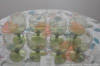 Front view of 8 avocado green footed glasses arranged in two rows on a floral tablecloth.