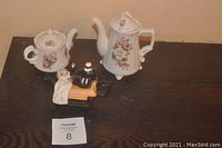 Three single serve decorative teapots placed together on a dark surface. Two are white with floral designs, and one is shaped like a vintage sewing machine.