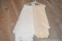 Photo of two folded lace tablecloths on a hanger, one white with intricate lace edges and one beige with a waffle weave pattern.