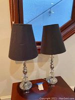 Pair of chrome and glass table lamps on a wooden surface with black shades, positioned near a window with daylight.