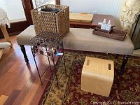 Wide view of upholstered bench with cushions, plant stand, wicker basket, step stool and trays.
