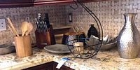 Photo shows entire kitchen corner countertop with wooden utensil holder, Sabatier knife block, decorative bowls, wire fruit basket, decorative platter, hammered vase, and paper towel holders.