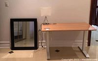 Wooden desk with white metal legs, silver table lamp with square lampshade on desk, black framed mirror leaning against wall on floor.