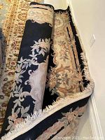 Partially rolled large floral pattern floor rug with black base and beige floral border with tassel fringe.