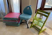 Blue kids office swivel chair, green Stokke high chair, and stack of foam puzzle mats clustered near window.
