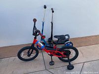 Red Kid's Trek bicycle with training wheels and 2 kick scooters behind it against white wall and concrete ground.