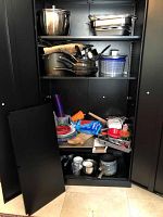 Metal cabinet with door removed showing kitchen items inside including pots, pans, plastic dinnerware, utensils, and more.