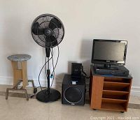 Photo showing black standing air fan, wooden stool, Polk Audio subwoofer speaker, Philips HDTV, Netgear router on wooden side table with shelves