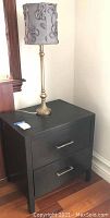 Full view of black two-drawer nightstand with gold table lamp on top, featuring gray lamp shade with ribbon design.