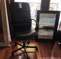 Black adjustable office chair with armrests and caster wheels next to a rectangular wall mirror with a thick black frame placed on a wooden floor.