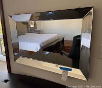 Front angle view of wall hanging beveled mirror mounted on wall reflecting chair and bed