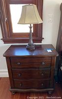 Wooden nightstand with three drawers and a table lamp on top, shown next to a window with wooden frame.