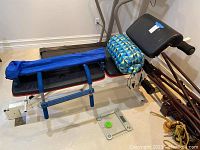 Workout bench with attached foam-padded pull-up bar and rectangular padded backrest. Blue geometric patterned Coleman sleeping bag in a bag lies across the bench. Digital glass weight scale on the floor.